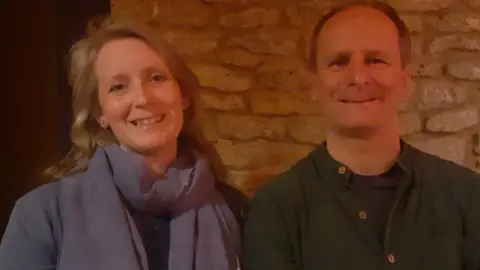 Picture of Mel Dalton and Neil Croom smiling beside each other at a meeting of Grief Network. Mel is wearing a purple top and a purple scarf and Neil is wearing a dark-coloured T-shirt under a shirt.