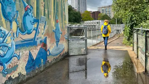A woman in a yellow rain coat and blue trousers is walking on a bridge over the river. There is a colourful mural of blue characters on the wall in the foreground. The woman is reflected in a puddle on the path. In the background you can see trees and tall buildings. 