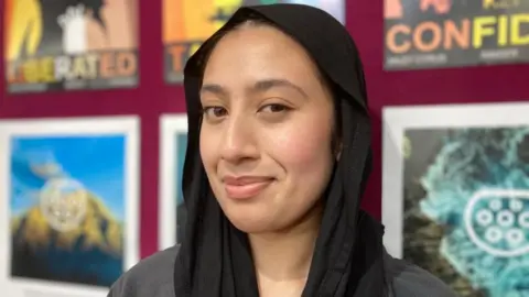 Alex Pope/BBC Fatima, wearing a black scarf on her head, looking straight at the camera, standing by a wall with artwork on behind her.