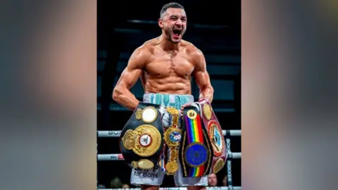 Boxxer Callum Simpson in the boxing ring. He is holding four belts across his front and shouting in celebration. He is muscular and wears long white boxing shorts.