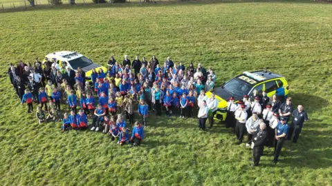 Wiltshire Police A drone shot of more than 100 children looking upwards in guides and brownies uniform. Next to them are police officers and two police cars in a green field.