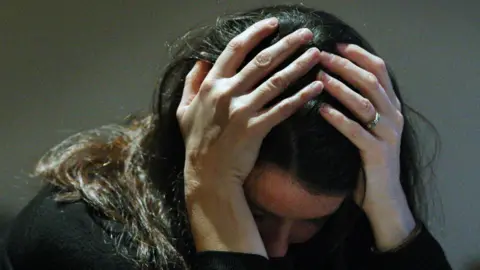 A picture of a woman with her hands in her head. She has long black hair and is looking down.