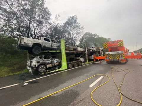 Leicestershire Fire and Rescue Service Multiple burnt out vehicles are held on a car transporter on the M1. An orange recovery vehicle is parked on the carriageway next to the transporter.