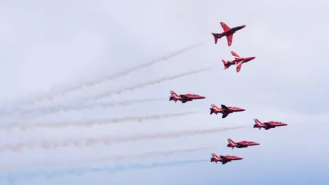 PA Media Seven red arrows in the sky with white and blue smoke coming out from behind them. Blue and white skies behind.