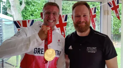 Supplied A picture of Aaron Phipps in a Great Britain tracksuit holding his gold medal and Jon Cooper standing in front of Union Jacks in front of a garden. 