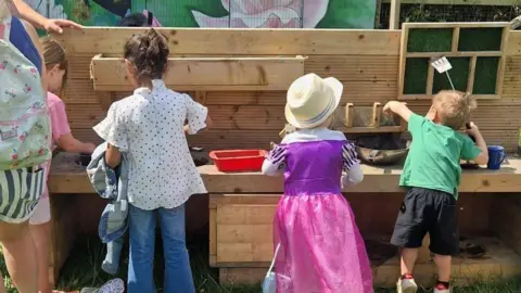 The Green Backyard Four children- playing on the kitchen garden- placed within a wooden frame- placed on the grass- with their backs to the camera