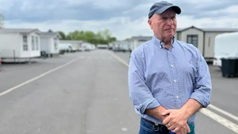 Billy Welch, wearing a baseball cap, blue shirt and jeans, gazes into the distance with his hands clasped in front of him. He's standing in the middle of a road that cuts through his caravan site. Behind him are rows of caravans.