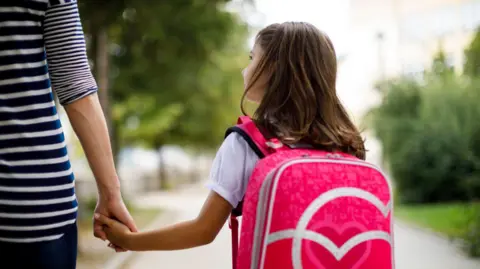 The back of a young girl, with long brown hair, wearing a white t-shirt and pink backpack. She is holding a woman's hand. 