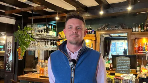 Ian Hillman wears a blue gilet and stands in front of the bar at his pub, a Sixteenth Century inn. He has a goatee, and is smiling. 