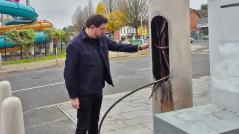Paul Doherty A man is standing next to a damaged mast. A leisure centre and other businesses and shops are in the background. The door of the mast is open and the wires inside have been burnt.