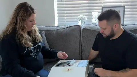 Shane Bevan and Laura Tongue looking through a memory box after the death of their son. They are sitting ona grey sofa in their home