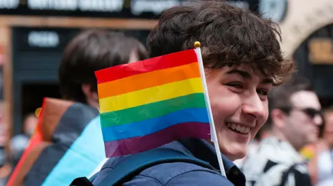 A Pride parade attendee looks toward the camera while holding a small rainbow flag. Other participants and city buildings appear blurred in the background.