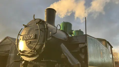 A green and black steam locomotive lit up by the late afternoon sun. The badge on the front bears the number 30075. Steam is coming from the chimney on the top.