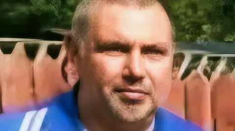 Cornelius O'Neill has short hair and is wearing a blue shirt. He is standing near a fence. 