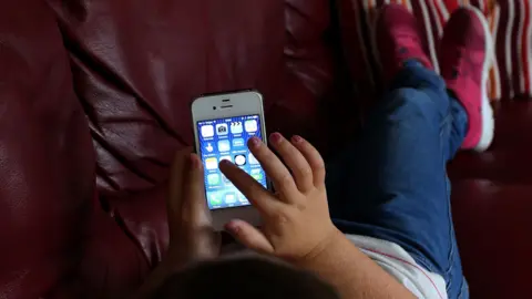 PA Media A child lies on a red sofa and is scrolling along a mobile phone screen with their finger