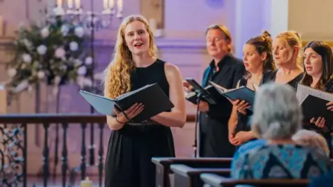 Classical singer Lucy Cox is singing in a choir dressed in black, with other singers stood behind her and to the right.