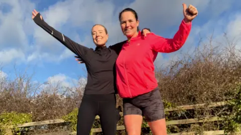 Michelle Kelly Sisters Michelle Kelly and Emma dressed in running clothing. Both are smiling looking straight at the camera with their arms stretched wide. 