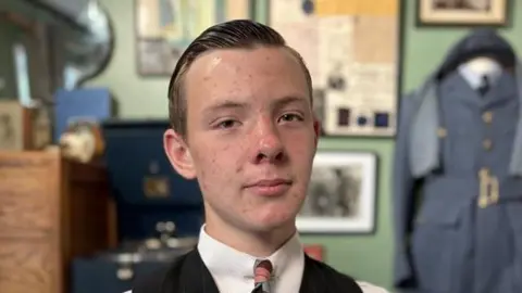 Lincoln smiles at the camera in his bedroom that is decorated in 1940s memorabilia. He has short hair that has been jelled back. He wears a white shirt, tie and black waistcoast. 