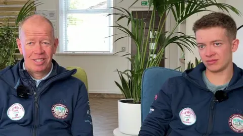 Paul and Aaron Anderson wearing Everest Summit Challenge 2024 badges sitting in room in front of two plants 