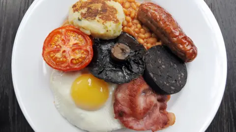PA Media A white plate with the items that make up a full English breakfast laid out on it.