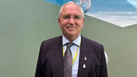 Police and Crime Commissioner Chris Nelson is dressed in a dark suit and stands in front of the mural which features an olive green base and a daylight skyline above him.