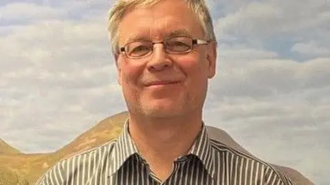 A grey-haired man with glasses and striped grey shirt looks at the camera