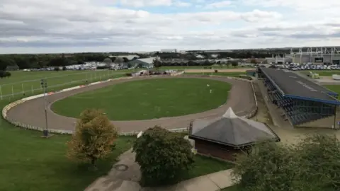 Steve Hubbard/BBC Aerial view of the speedway track on the showground, with spectator stand to the right and buildings in the distance. 