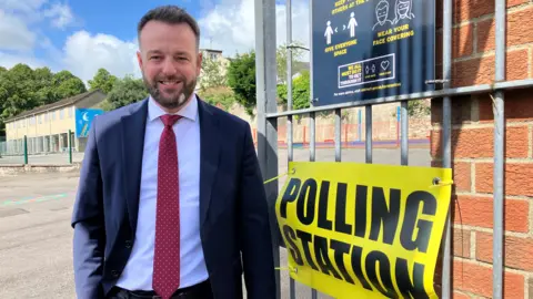 Man in blue suit, white shirt and red tie with a beard outside a polling station