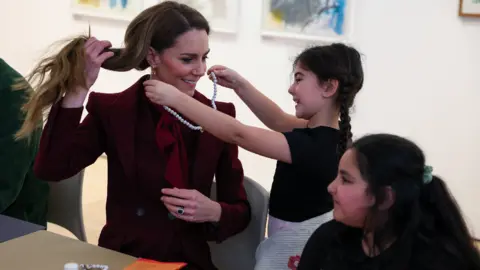 PA Media A girl holds up a necklace around the Princess of Wales' neck, while another girl looks on