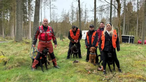 Group of people wearing high-visibility orange vests standing in woodland with their dogs.