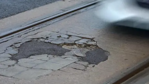A badly damaged and cracked road surface, with tram tracks on either side.