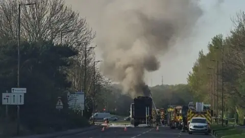 HIWFRS Large lorry on fire with vast black plume of smoke coming from it. Multiple fire engines can be seen alongside the lorry.