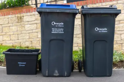 Wiltshire Council Three bins lined up next to each other on a pavment. They say 'recycle for Wiltshire' on them
