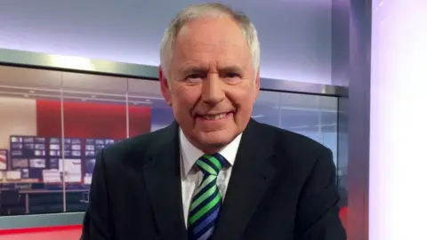 Nick Owen is sitting in a TV studio looking at the camera. He is wearing a dark jacket, white shirt and green and blue striped tie.