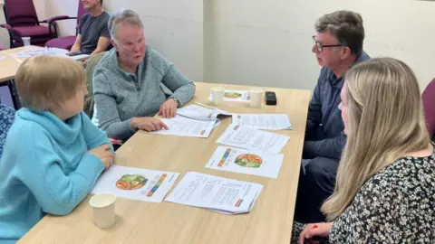 Four people sit around a wooden table with leaflets in front of them. One of them is a blonde haired younger woman who is a health coach and three others are slightly older people, two woman and one man.