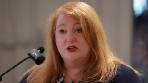 Naomi Long is standing in front of a microphone, She has ginger hair and is wearing red lipstick. 
