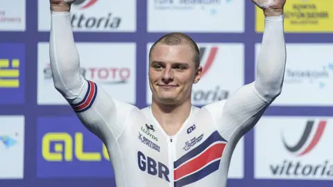 Matt Richardson in white British kit with GBR lettering raises arms aloft in front of sponsor background