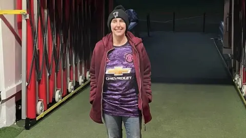 Zoe Hodges Zoe wearing a purple Manchester United shirt, a burgundy hoodie, black beanie and jeans facing the camera while stood in the tunnel at Manchester United's ground, Old Trafford.