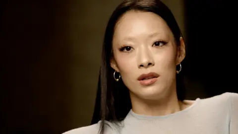 Rina Sawayama sitting for interview with BBC, she wears a light blue top and hoop earrings.