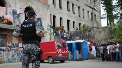 BBC A police officer in a stab vest watches a long line of men on the stairway into the shell of a building