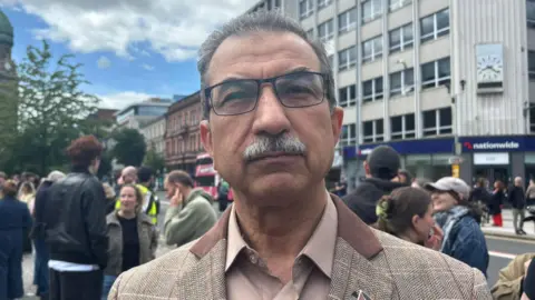 Raied is looking straight astatine the camera. He is wearing a tweed blazer and a nude coloured shirt. He has grey hairsbreadth and square-framed glasses.