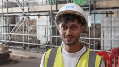 Samuel Javid smiles at the camera as he stands inside the Jacob Wells Baths. He is wearing a hard hat, with shirt and a high-visibility jacket. There is scaffolding in the background.