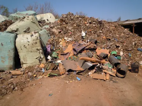 Environment Agency A pile of decomposing waste which had been dumped at a site in Lincolnshire. Blue bales are stacked at the left of the image but have partially split. A large mound of brown-coloured rubbish, including what looks like pieces of metal, is visible on the right of the image.