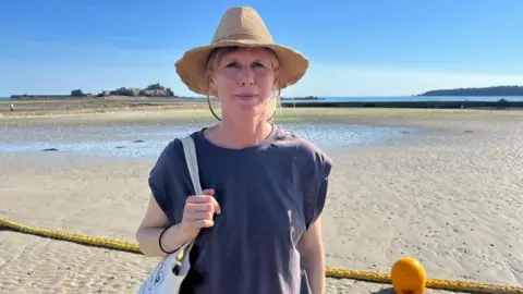 A white woman wears a light brown summer hat. She wears a navy t shirt and holds a white tote bag on one shoulder. She's standing at the beach, with Elizabeth Castle in the background. It's low tide. Behind her is a yellow rope and buoy on the sand.
