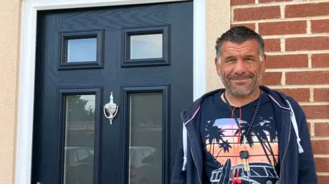 JOANNA TAYLOR/BBC Paul Smith is standing smiling at the camera. He is wearing a T-shirt with a picture of palm trees on it and a navy hooded jacket. There are keys and earphones around his neck. Behind him is a black door with windows and a brick wall.
