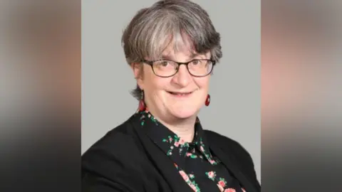 A woman with short black and grey hair looks into the camera, wearing black-rimmed glasses. She has a dark jacket on over a black shirt with a light floral pattern.