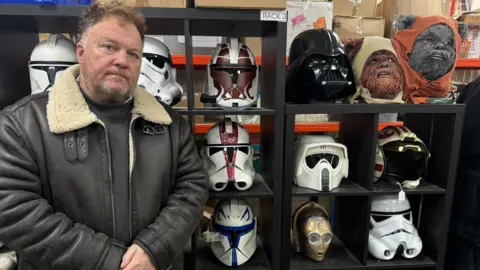 A man wearing a leather jacket stood in front of shelves. The man is looking at the camera. He has brown hair and a beard. On the shelves are helmets from the Star Wars films.