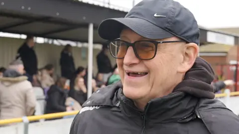 Connor Bennett/BBC Jim Purtill who is wearing a black baseball cap and black coat and black framed glasses. He is smiling broadly. Behind him is the stand with men and women standing and sitting. 