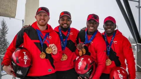 Antony Scully/Scully Photography Four Trinidad and Tobago athletes in red helmets and black‑and‑red winter sports gear stand together on snow, smiling and wearing multiple medals, with an ice track structure behind them.