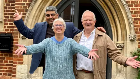Three people - two men and a woman - stood outside a church with their hands in the air in a celebratory way